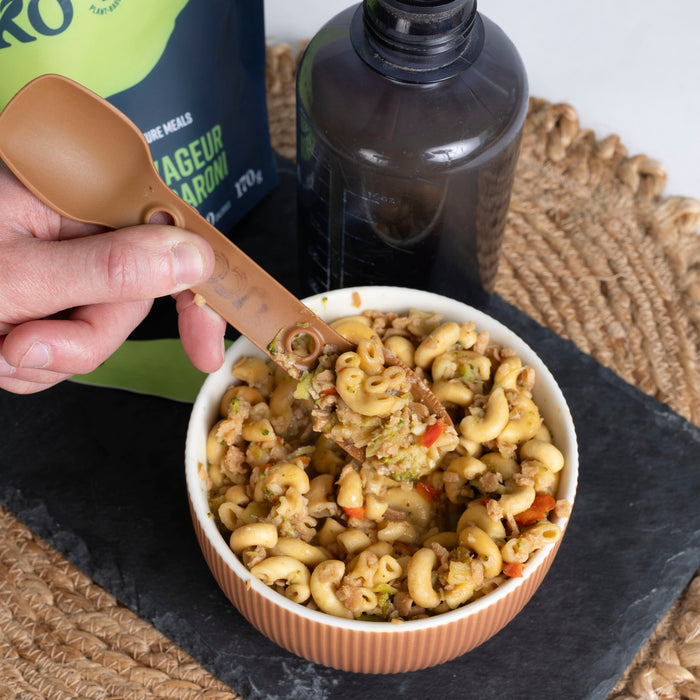 Person enjoying Treko Voyager's Macaroni in wooden bowl. Hand with fork poised over colorful pasta dish. Green packaging visible nearby.